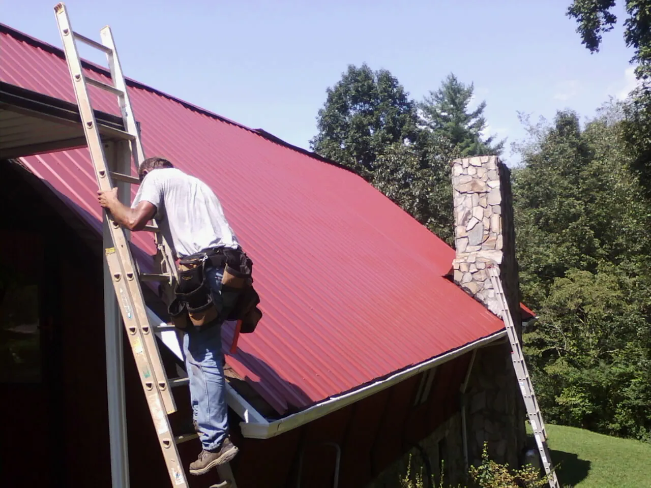 Roofer on a ladder beside a red metal roof and stone chimney — Soffit & Fascia Repair by Roofing Brothers in Liberty Lake